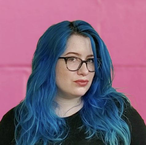 A woman with blue hair and glasses in front of a pink wall.