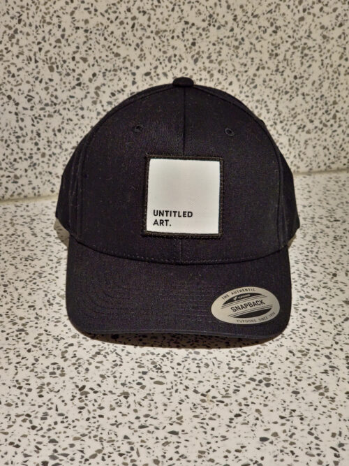 An Untitled Art Patch Logo Hat – Black with a white patch on it.