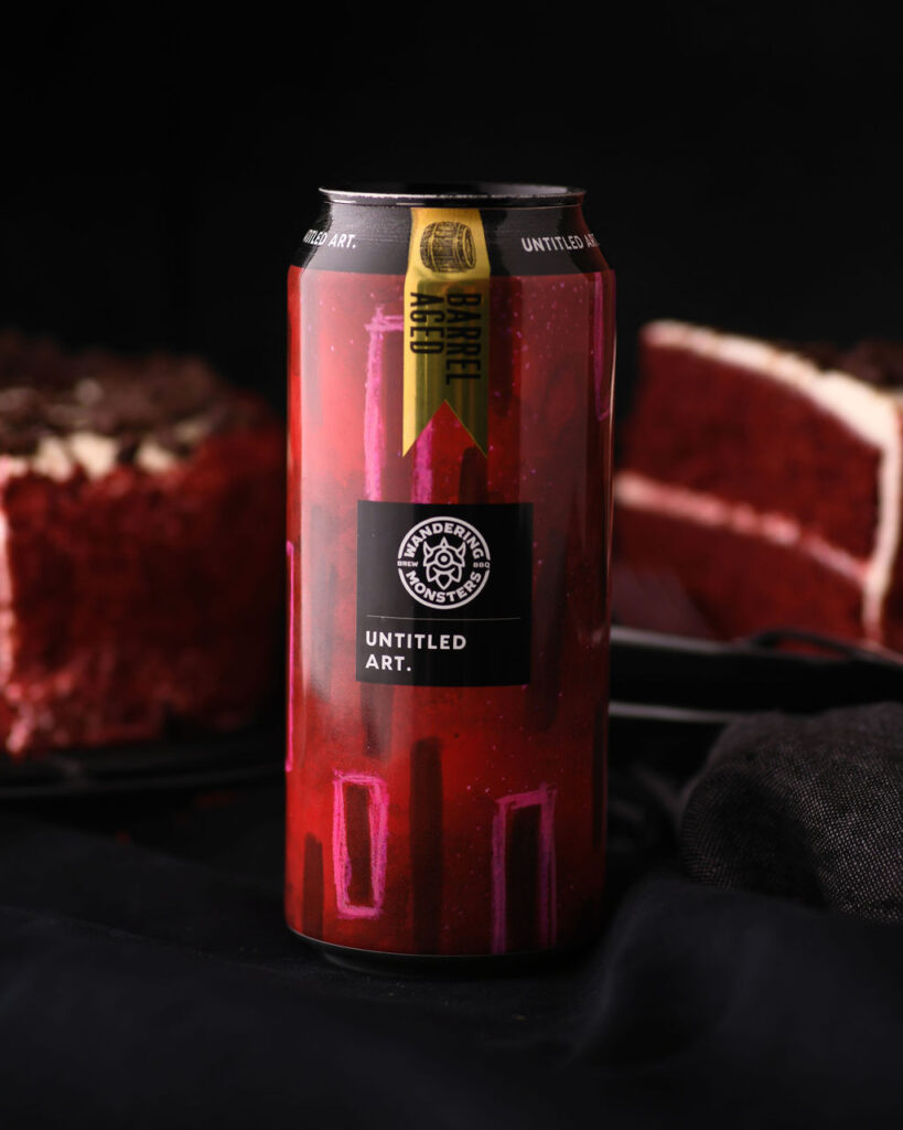 A can of untitled art beverage with a vibrant red design, placed in front of a red velvet cake on a black background.