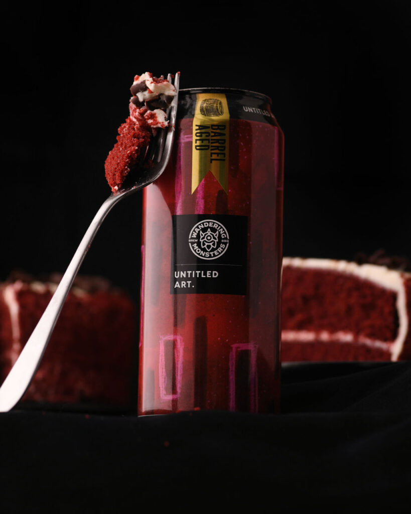 A piece of red velvet cake on a fork in front of a can of untitled art beverage with a cake backdrop.