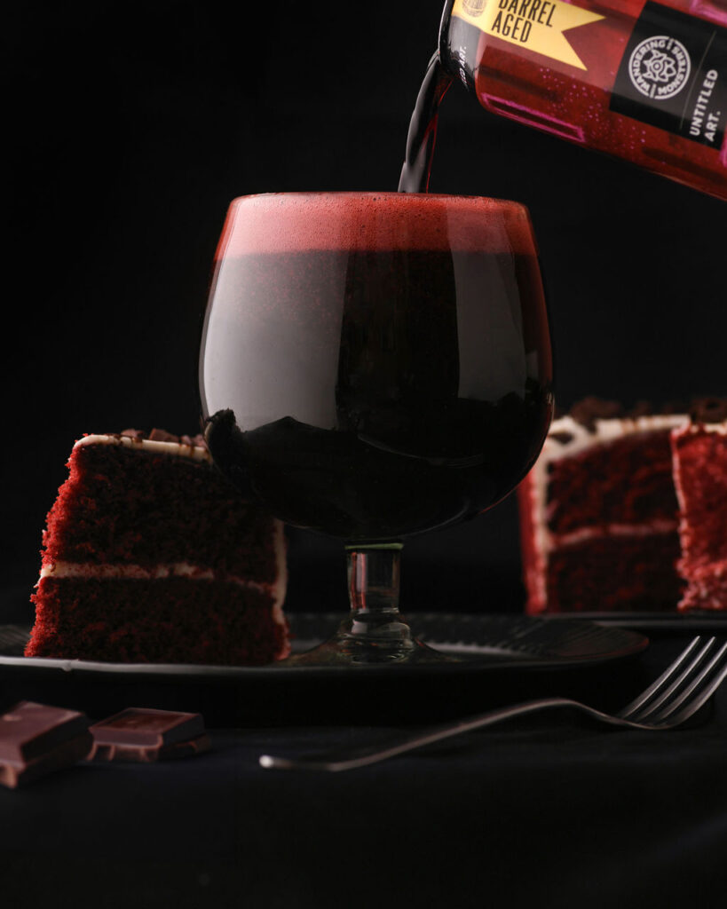 Dark beer being poured into a glass next to slices of red velvet cake against a black background.