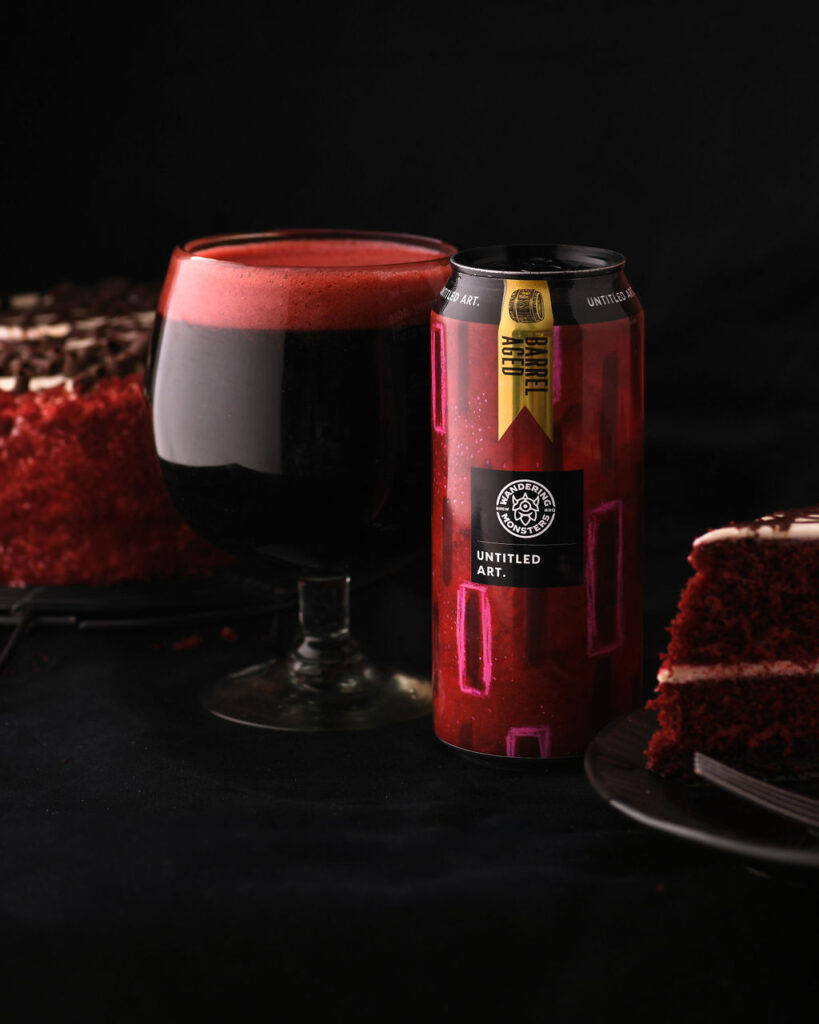 A can of untitled art beverage beside a glass of dark beer with a frothy head, set against a backdrop featuring a slice of red velvet cake.
