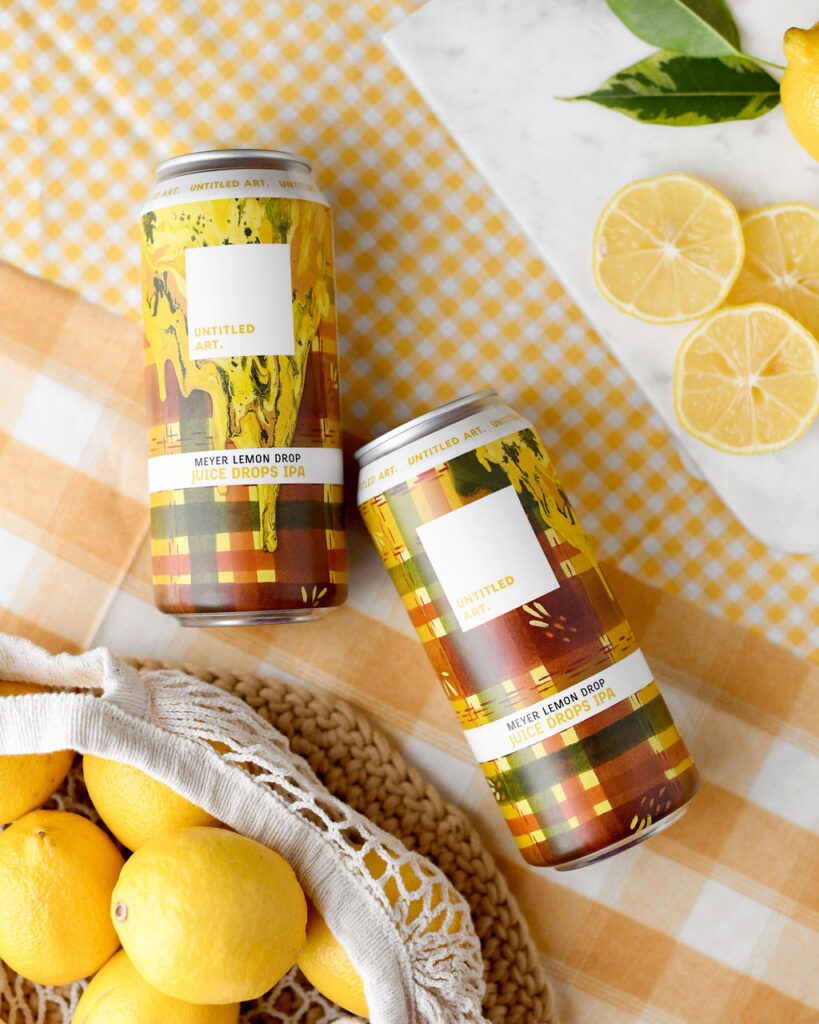 Two cans of meyer lemon drop juice on a table surrounded by fresh lemons and a checkered yellow cloth.