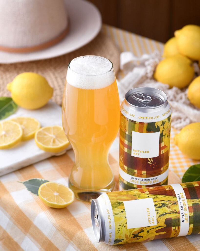 A glass of beer alongside a can with a lemon-themed label, surrounded by fresh lemons on a checkered tablecloth.